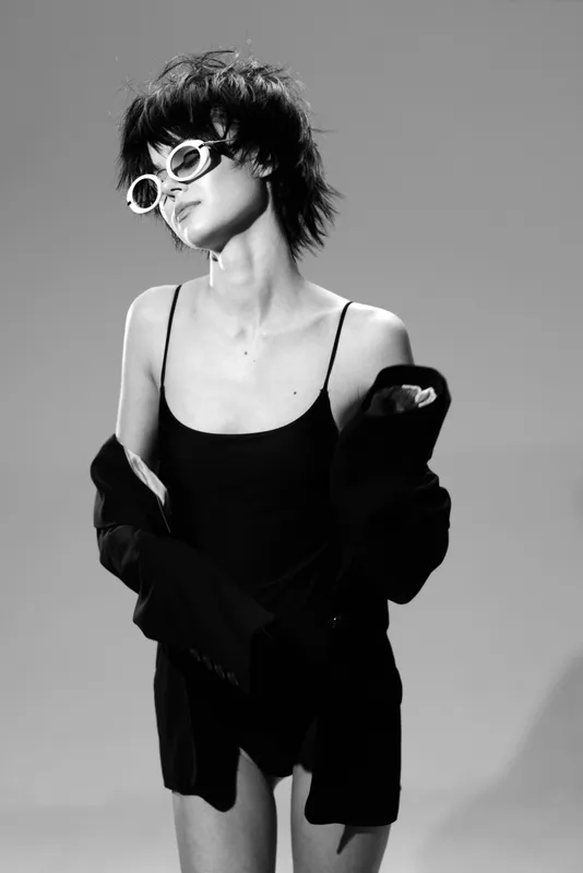 Black and white portrait of a stylish woman wearing sunglasses and a black outfit with an oversized jacket, showing confident and relaxed pose.