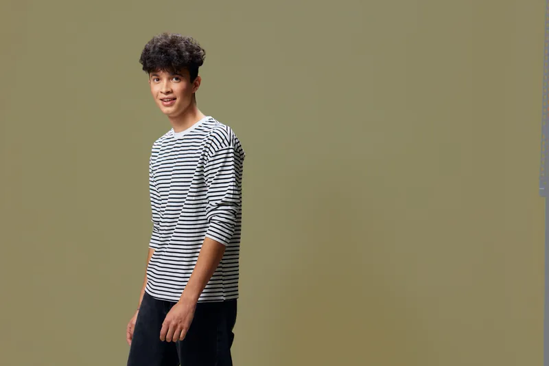 young man wearing striped shirt standing against plain background with casual posture, short curly hair, and relaxed expression in natural light