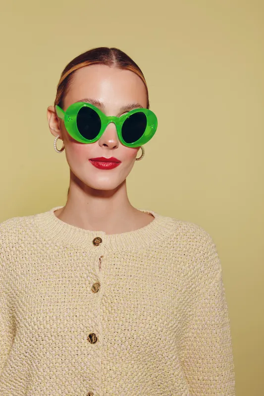 Young woman with slicked hair wears bright green round sunglasses and beige knit sweater against yellow background. Fashion portrait with bold accessories and minimal style.