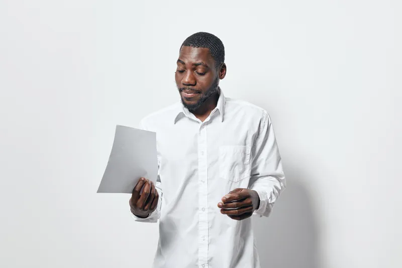 African American man with dark skin reading document in white shirt standing against white background. Serious expression, casual business style, isolated studio portrait with copy space.