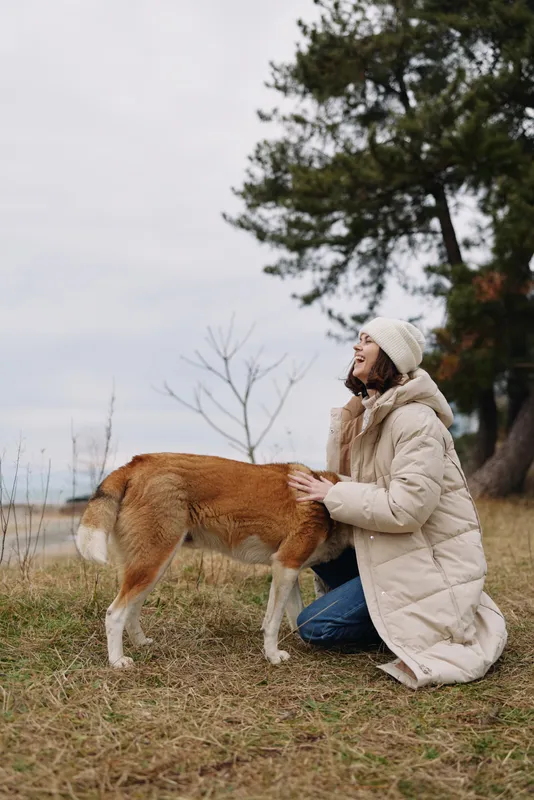 Woman kneels beside a dog in a chilly outdoor setting, wearing a thick coat and knit hat, sharing a quiet moment of companionship among tall trees and dry grass.