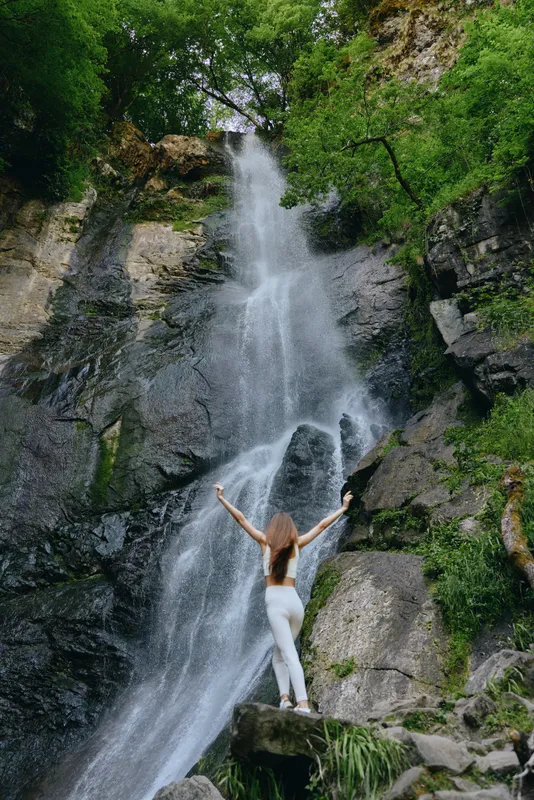 woman standing at the base of a tall waterfall in lush green forest, wearing white pants and raising her arms, enjoying nature and outdoor adventure.