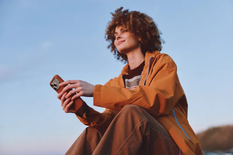 Woman lifestyle outdoors smartphone portrait sitting curly hair, warm field background, she holds smartphone with both hands, relaxed smile while sitting, casual autumn jacket, blue sky and natural