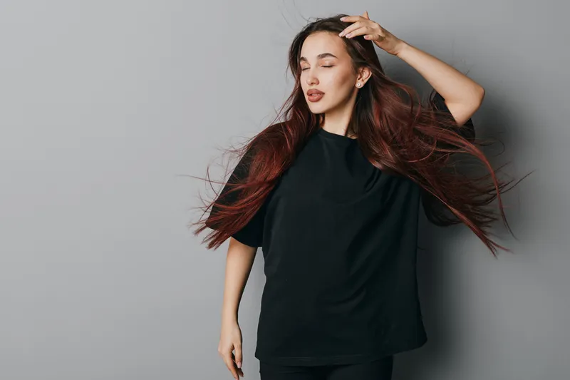 Woman with long hair wearing black shirt posing with closed eyes and hand on head against gray background. Fashion model expressing calm and relaxed mood with flowing hair.