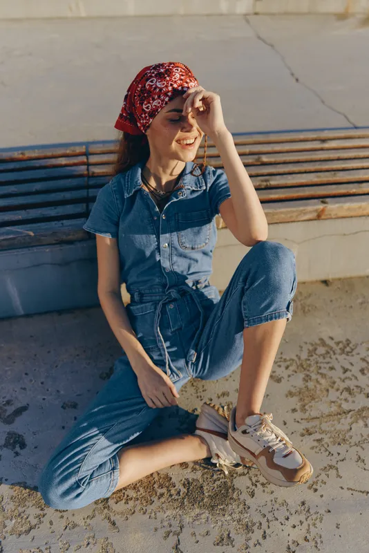 Lifestyle woman in modern boho-western denim street style wearing a red bandana, relaxed pose, natural sunlight, warm film color tone, casual outdoor setting.