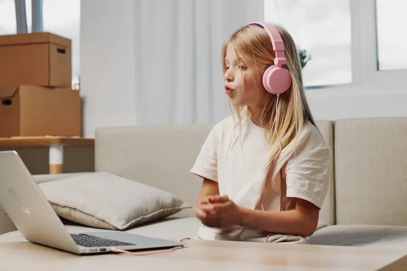 Young girl wearing pink headphones watches laptop screen attentively sitting on beige sofa in bright living room. Concept of remote learning, digital communication, and childhood technology use.