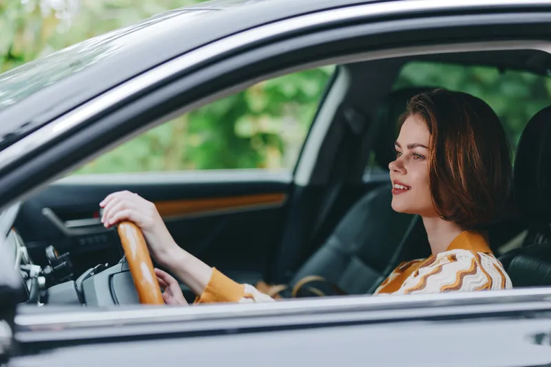 A woman is driving a car, hands on the steering wheel, looking ahead with a calm, focused expression inside a modern cabin.