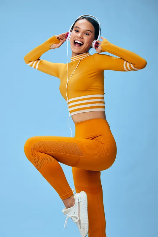Woman fitness portrait: energetic young woman in orange athleisure set and white headphones jumps mid-pose, sport concept emphasizing active lifestyle, sneakers and confident smile against blue