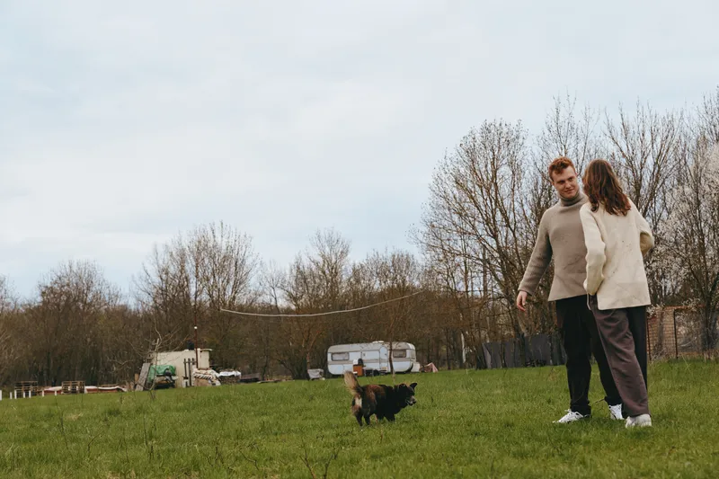 A couple stands in a grassy park as a playful goat kid approaches, capturing a relaxed lifestyle moment outdoors, highlighting bonding and gentle interaction in a natural setting