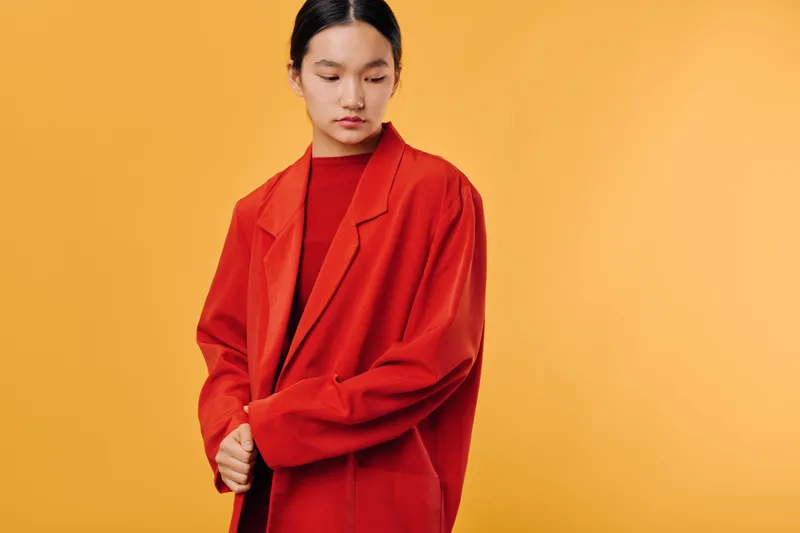 Young woman with long hair wearing red jacket and turtleneck posing against orange background with calm expression and casual style for fashion or portrait concept.