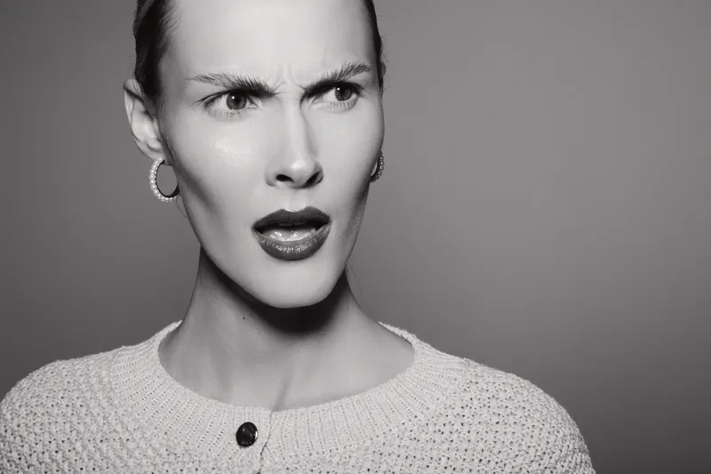 Confused white woman with fair skin wearing earrings and knitted sweater, showing disbelief or surprise on gray background.