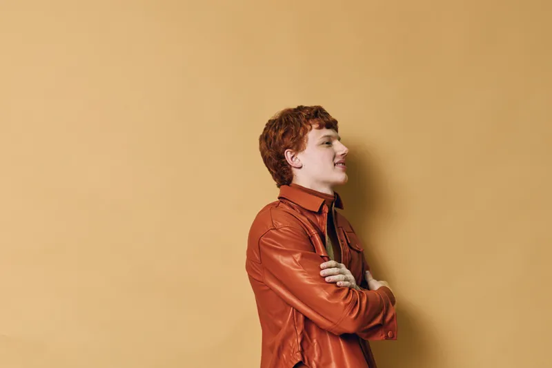Man with red hair wearing brown leather jacket stands with arms crossed against beige wall in casual pose looking sideways with confident and relaxed expression indoors.