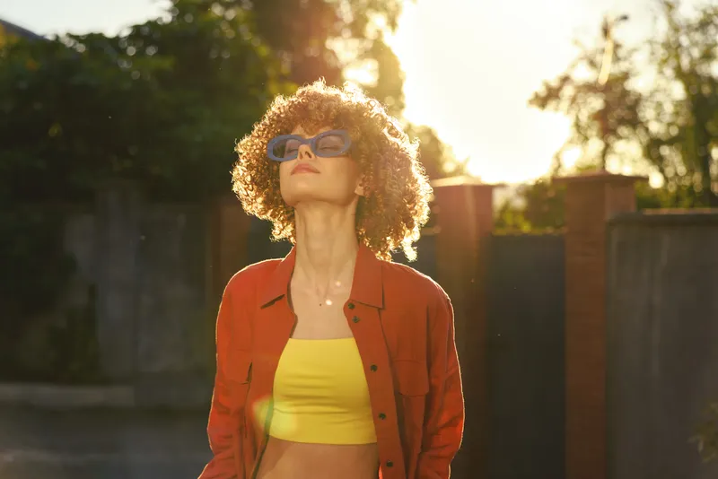 Cheerful woman outdoors, curly hair, yellow top under red jacket, sunlit moment, relaxed pose, natural colors, lush greenery in the background, gentle breeze, warm glow, casual style