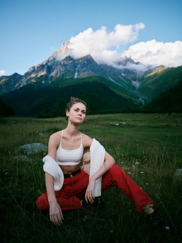 Woman sits in a grassy field with a mountain backdrop, wearing a white crop top and red pants, relaxed pose in natural light, outdoor scenery and serene alpine landscape