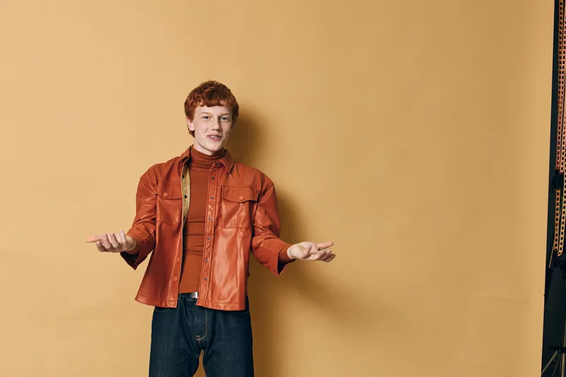 Young man in orange leather jacket and dark jeans posing with open hands against beige background studio portrait, fashion and casual style concept.