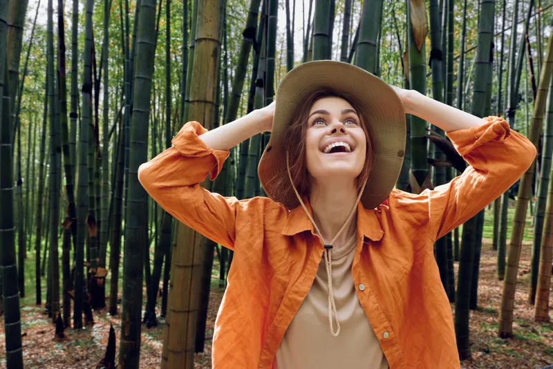 Woman in hat stands amid tall bamboo forest, smiling and looking up with joy. Outdoor portrait, orange jacket, nature travel, peaceful green grove and happy candid expression.