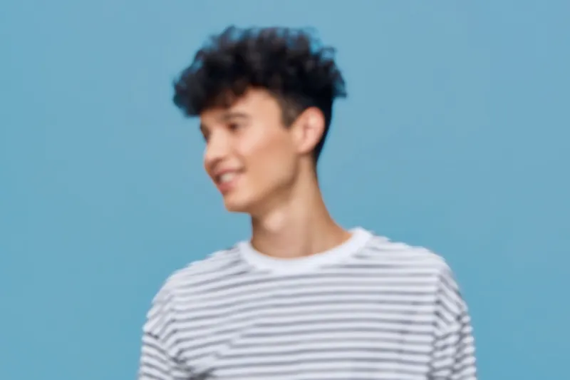 young man smiling happily wearing striped shirt with curly hair against soft blue background in casual style, creative portrait with natural emotion and relaxed posture