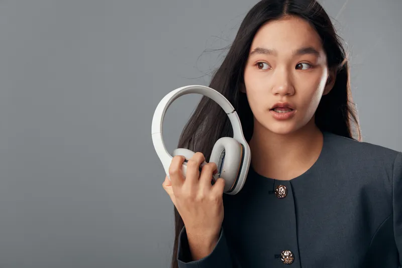 Woman holding white headphones with one hand looking to side wearing dark jacket on gray background. Female adult portrait with long hair and casual expression in studio environment.