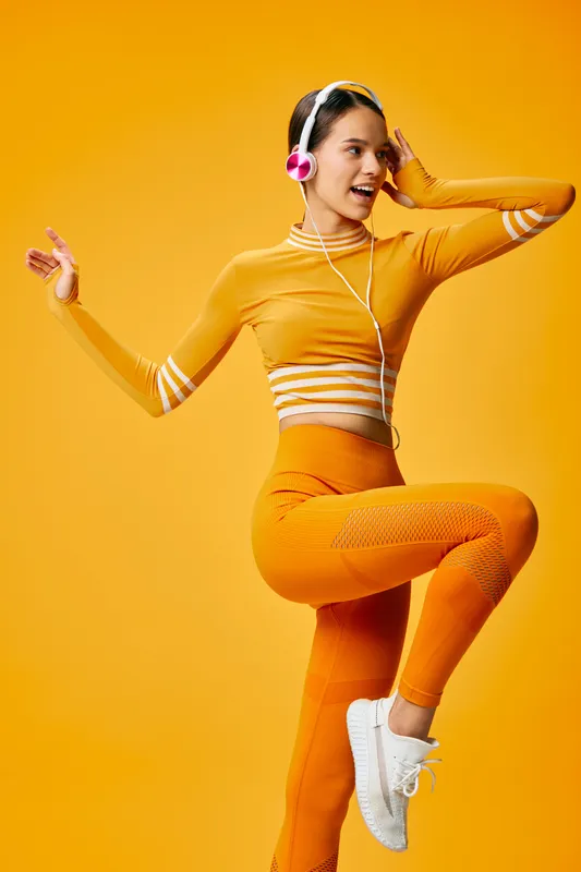 Energetic woman in orange athletic wear jumps with headphones on a vivid orange backdrop, showcasing fitness, dance and movement for dynamic sport lifestyle and active wellness.