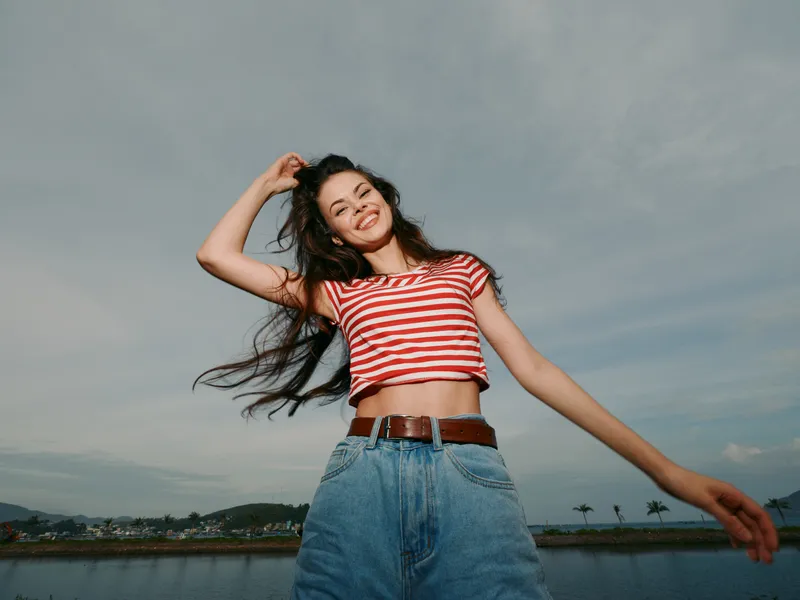 Young woman in a red and white striped crop top and high waisted jeans smiles joyfully outdoors by a calm waterfront, captured in bright daylight with carefree summer energy.