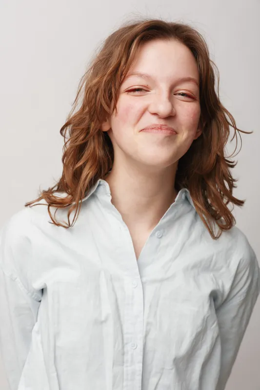 Smiling young woman with curly brown hair wearing white shirt stands against light background. Natural expression and casual style create friendly and relaxed mood in studio portrait.
