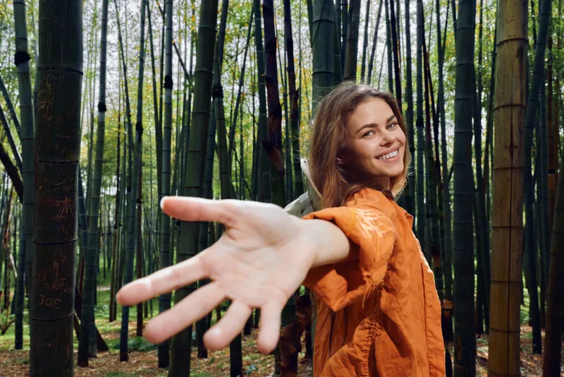 Woman smile hand reaching forward in bamboo forest, portrait of happy traveler in orange jacket inviting connection and adventure, eco nature scene with warm expression.