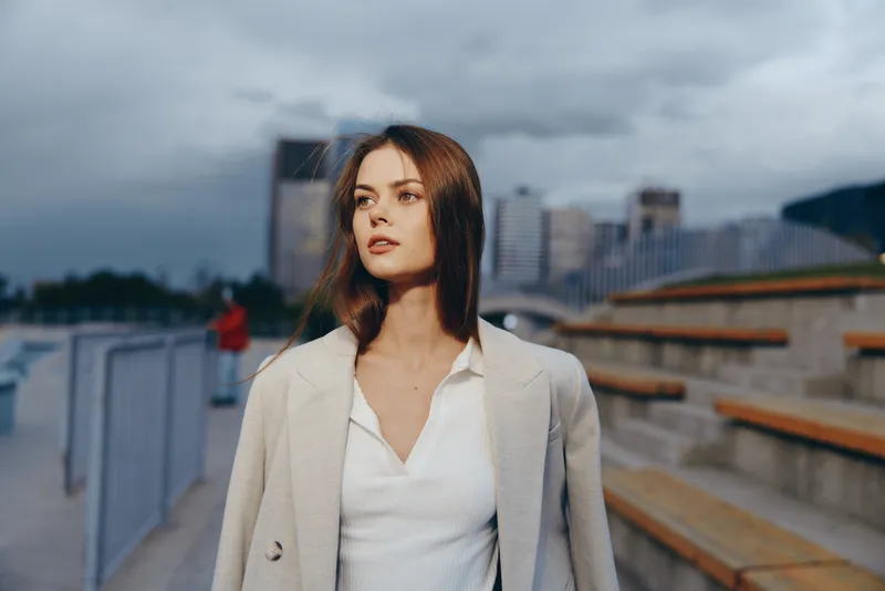 Young European woman in light coat with thoughtful expression outdoors in urban setting with cloudy sky and modern buildings, fashion concept