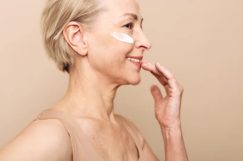 Smiling mature woman applies white face cream on cheek, skincare concept for aging skin. Soft light highlights natural beauty and self-care routine on beige background.