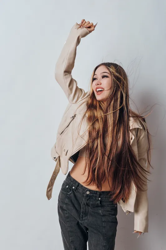 Young woman with long hair wearing light jacket and black jeans poses with raised arm and smiling face against plain background in studio. Casual style and joyful expression.
