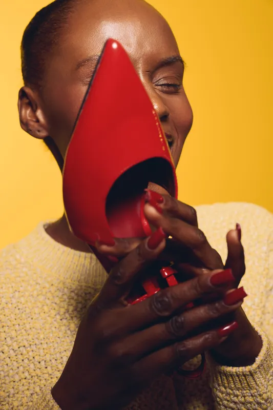Smiling black woman with dark skin holding a red high heel shoe near her face on yellow background. Concept of fashion, style, beauty, and joyful lifestyle.