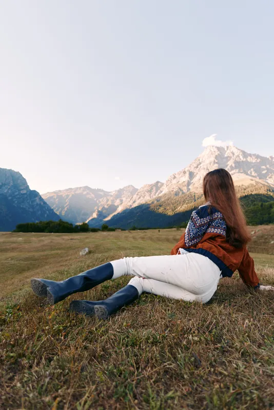 Woman relaxing in a meadow facing mountains and wide landscape, enjoying nature and quiet solitude. Casual sweater and boots, peaceful outdoor scene with scenic sunset light.