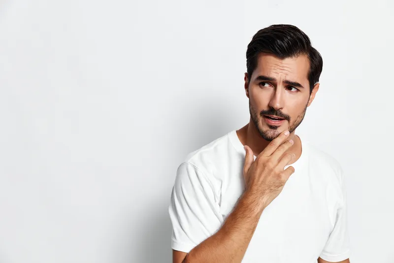 Thoughtful young man in white t-shirt touching his beard, pondering, looking sideways with a curious expression against a plain solid background. Lifestyle concept.