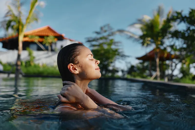 Calm woman rises from a pool with closed eyes, basking in warm tropical sun. A luxurious resort setting conveys wellness, relaxation, and selfcare through an outdoor lifestyle moment.