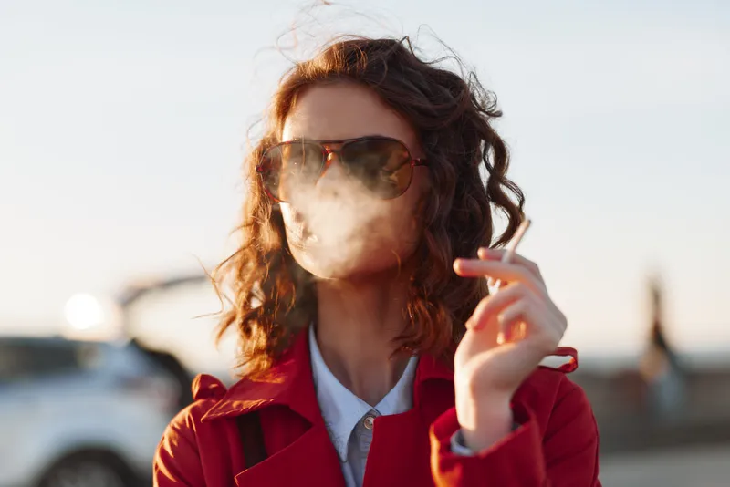 A young woman with curly brunette hair wears a red trench coat and oversized sunglasses exhaling smoke on an urban street for a bold, moody, cinematic moment