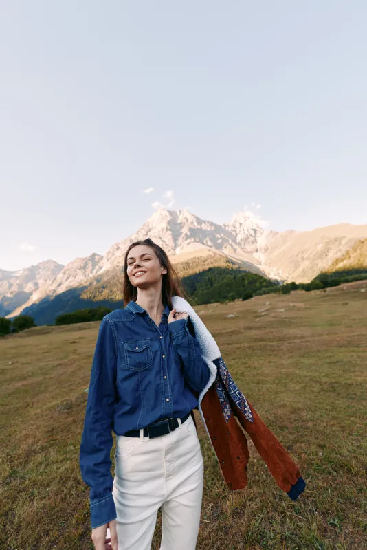 Woman smiling in mountains meadow wearing denim shirt and jacket, outdoors landscape scene of happy fashion portrait with casual clothing, sunlight, grass and serene alpine view.