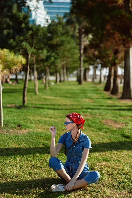 Lifestyle woman in modern boho-western denim street style sits cross-legged on grass wearing sunglasses and a red bandana, captured with warm film color tones.