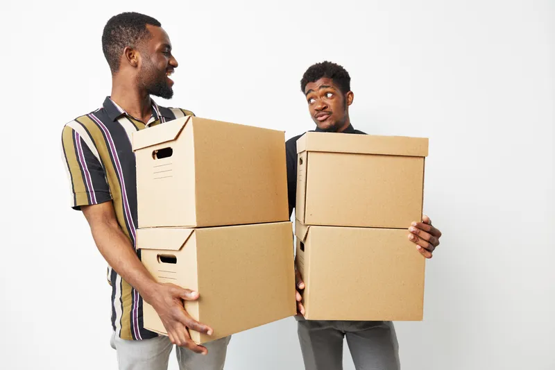 Two African American men smiling while carrying cardboard boxes, showcasing teamwork and camaraderie in a bright and minimalistic setting.