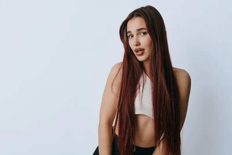 Young woman with long straight hair wearing white crop top posing against plain background. Female model with natural makeup and casual style looking at camera with relaxed expression.