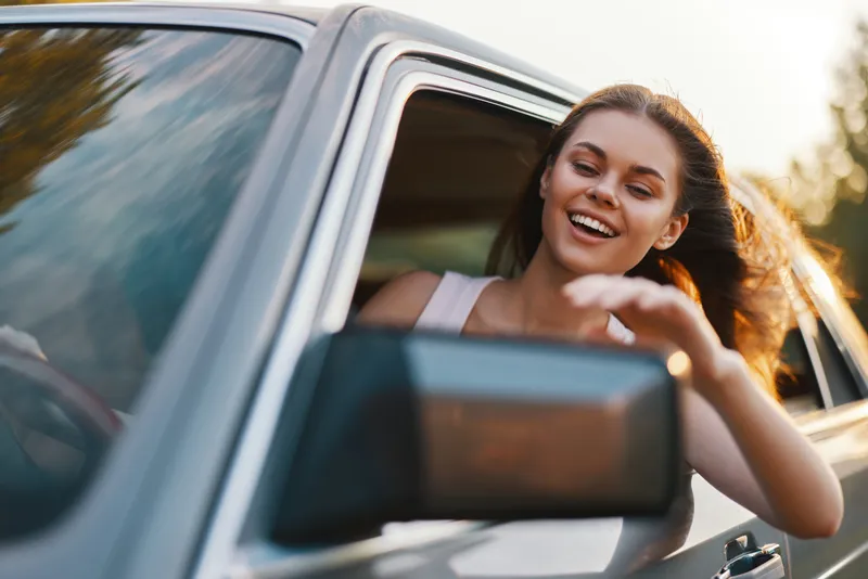 Happy young woman enjoying a road trip, smiling and waving from a car window, surrounded by a sunny backdrop, embodying freedom and adventure.