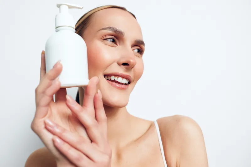 Smiling young woman with bare shoulders holds a white lotion bottle near her face against a plain white background, expressing skincare and beauty care.