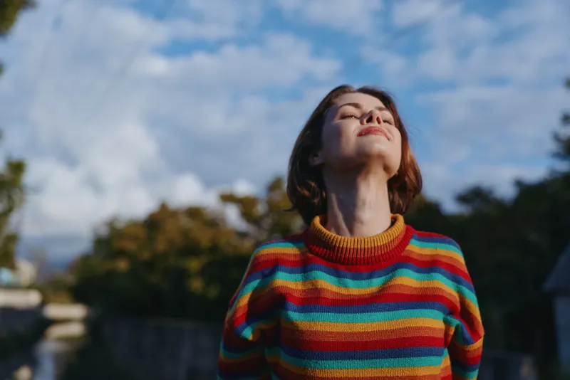 Woman portrait in colorful striped sweater outdoors under blue sky in autumn, relaxed and serene with closed eyes, enjoying sunlight, nature, fresh air and peaceful moment.