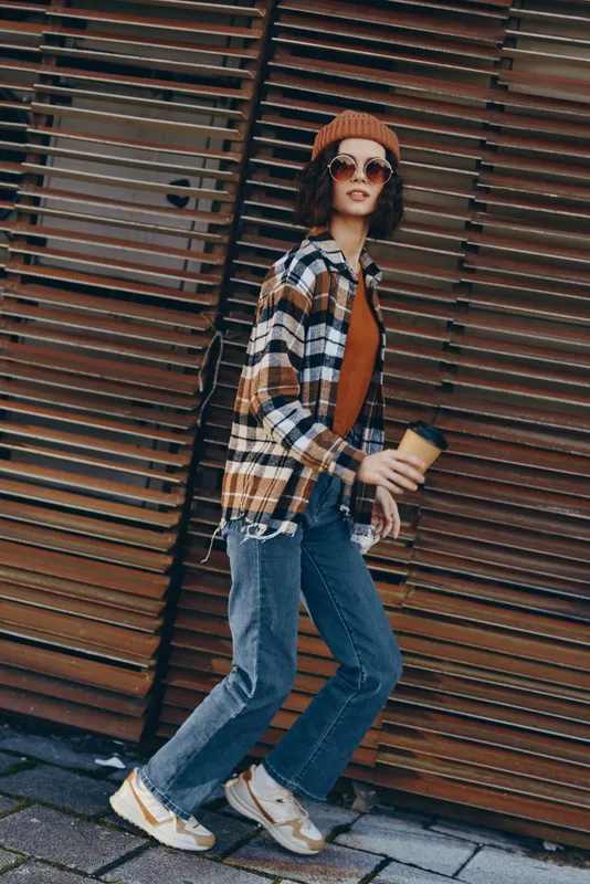 Woman walking urban street with coffee, wearing casual plaid shirt, beanie and sunglasses. Candid lifestyle portrait with authenticity, golden hour glow, mindful living and emotional storytelling.