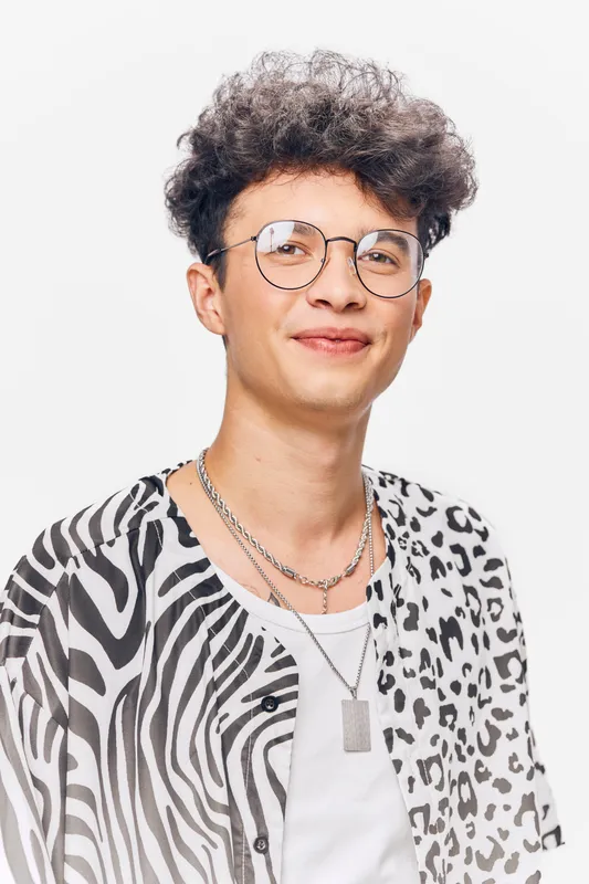 Young man smiling with curly hair and glasses wearing a mixed animal print shirt and layered silver necklaces against a white background.
