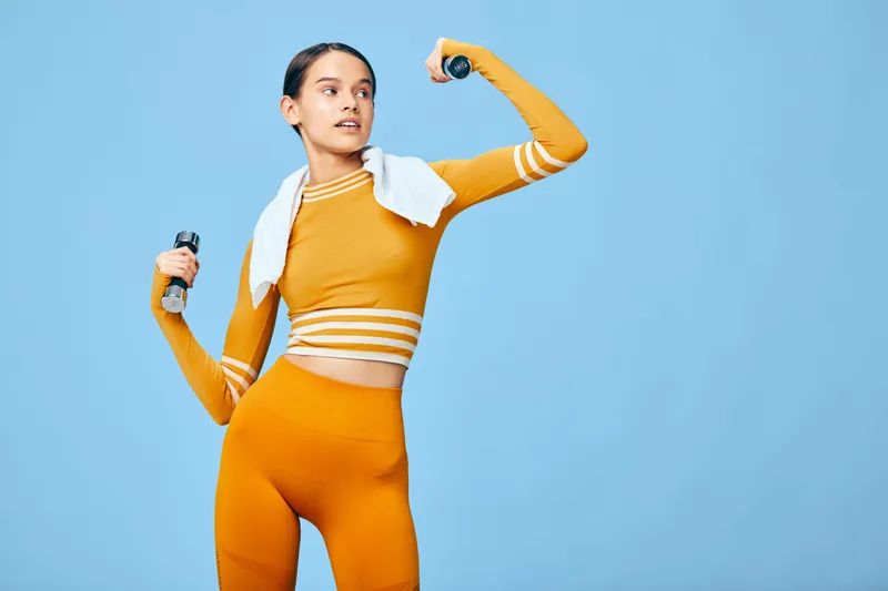 Fitness woman in bright orange athletic wear lifts dumbbells with a towel draped over her shoulders, performing a high energy workout against a vivid blue studio backdrop.
