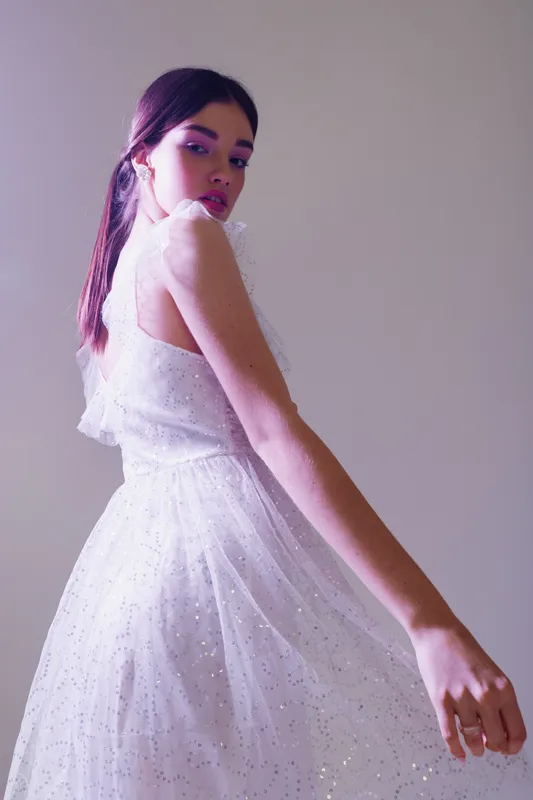 Young woman in a white sparkling dress poses with elegance against a neutral background. Soft lighting highlights her beauty and fashion style. Concept for beauty, fashion, and elegance.