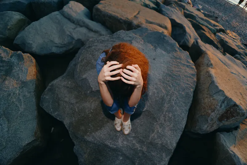 lifestyle woman with red curly hair in modern boho-western denim winter street style, film color, sitting on large rocks with hands covering her face