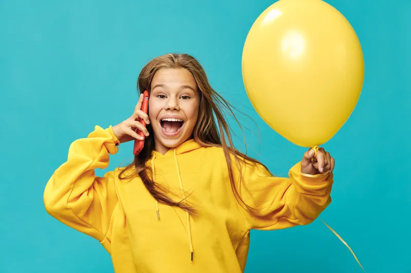 Girl in yellow hoodie smiling and holding yellow balloon talking on phone with happy expression on blue background bright casual style portrait
