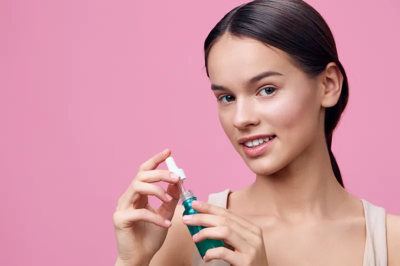 Young woman holding a serum bottle with a cheerful expression against a pink background, showcasing skincare, beauty routine, and healthy skin