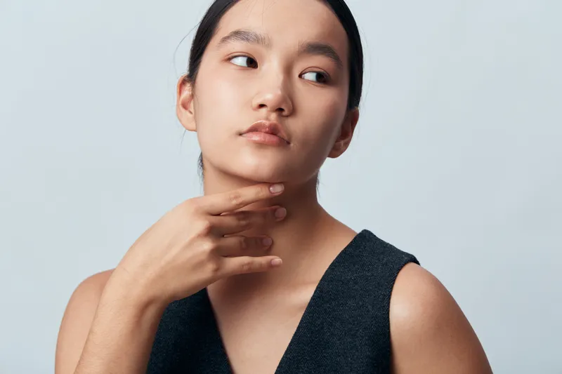 Young woman with long black hair wearing dark sleeveless top posing and touching her neck with thoughtful expression on light gray background in studio portrait.