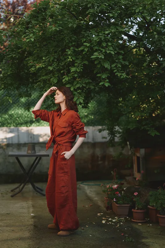 Woman wearing rust jumpsuit stands outdoors under a leafy tree, posing with one hand on her forehead, embodying casual chic lifestyle in a garden setting